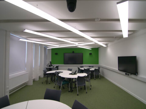 Shows one learning space with tables and a green wall with a big screen